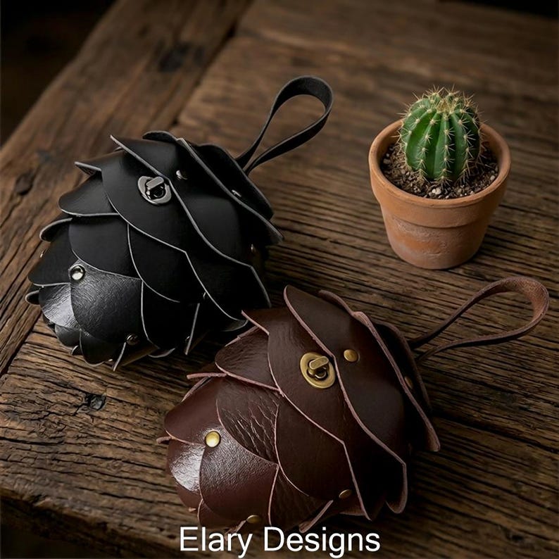 May include: Two leather keychains, one black and one brown, with a unique layered design resembling a pinecone. Each keychain has a small loop handle. A small potted cactus sits nearby. The text "Elary Designs" is at the bottom.