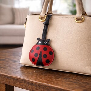 May include: A red and black ladybug-shaped bag charm with black spots and a black strap is attached to a beige handbag. The handbag has gold-colored hardware and is sitting on a wooden surface.