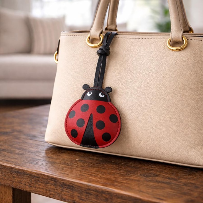 May include: A red and black ladybug-shaped bag charm with black spots and a black strap is attached to a beige handbag. The handbag has gold-colored hardware and is sitting on a wooden surface.