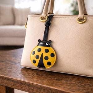 May include: A yellow and black ladybug-shaped bag charm with a black strap is attached to a beige handbag. The ladybug charm has black spots and a black head with white eyes. The handbag sits on a wooden table.