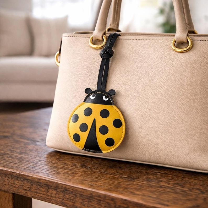 May include: A yellow and black ladybug-shaped bag charm with a black strap is attached to a beige handbag. The ladybug charm has black spots and a black head with white eyes. The handbag sits on a wooden table.