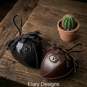 May include: Two acorn-shaped leather pouches, one black and one brown, with a handle and clasp. A small cactus in a terracotta pot sits between them on a wooden surface. The text "Elary Designs" is at the bottom.