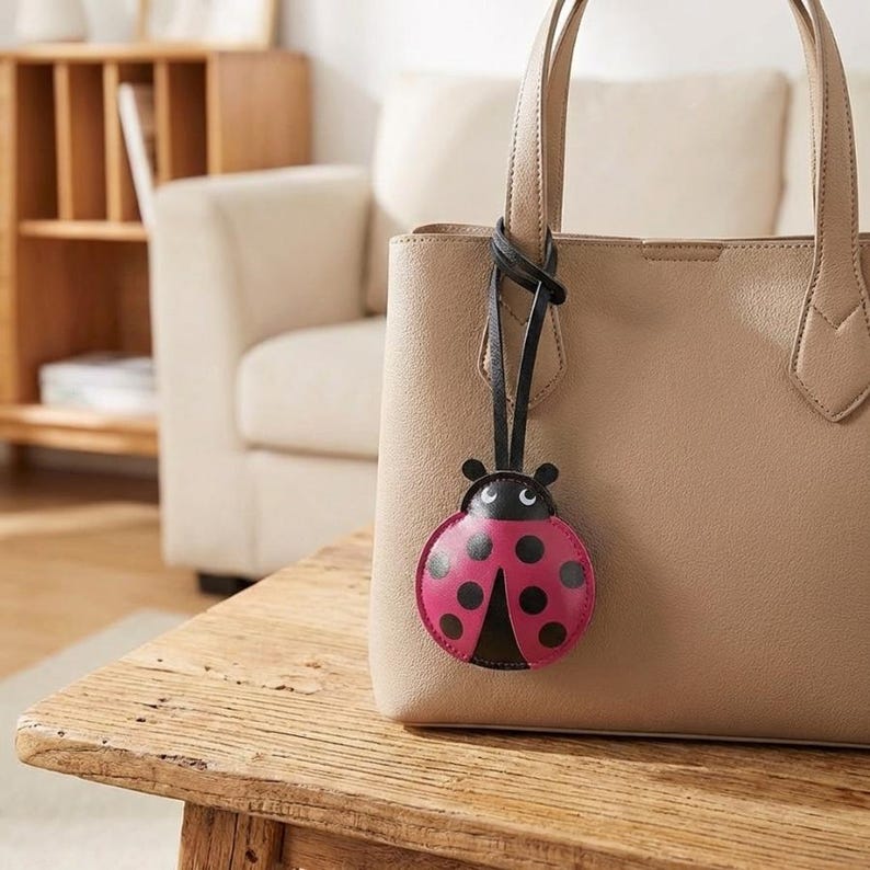 May include: A beige handbag with a black leather strap and a ladybug-shaped charm. The ladybug charm is pink with black spots and a black head. The bag is sitting on a wooden table.