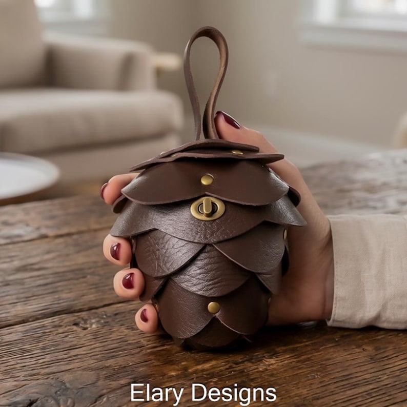 May include: A brown leather pinecone-shaped pouch with a handle. The pouch is made of layered leather scales and features a brass clasp. The image includes the text "Elary Designs".