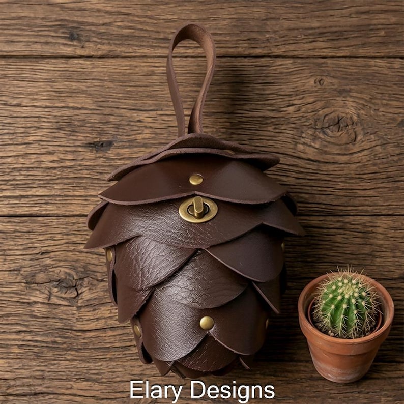 May include: A brown leather pouch shaped like a pinecone, with a handle and a clasp. The pouch is made of overlapping, petal-like pieces and secured with brass-colored rivets. A small cactus in a terracotta pot sits nearby. The text "Elary Designs" is visible.
