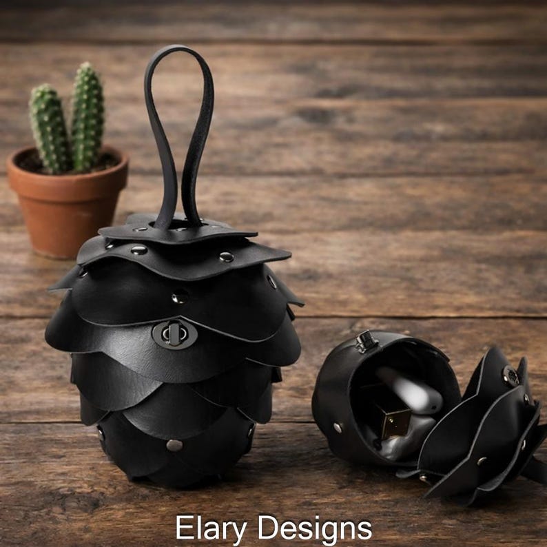 May include: Black leather pouch with a unique layered design, resembling a pinecone. The pouch has a handle and a clasp closure. The open pouch reveals small items inside. The text "Elary Designs" is at the bottom.