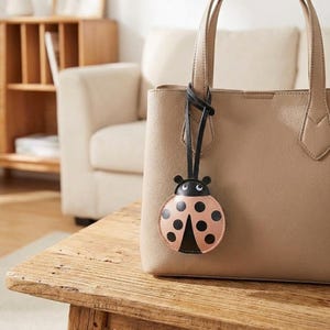 May include: A beige tote bag with a black leather strap and a ladybug-shaped charm. The ladybug charm is pink with black spots and a black head. The bag is sitting on a wooden table.