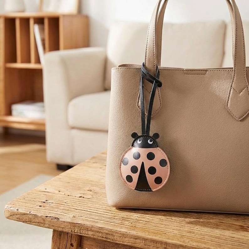 May include: A beige tote bag with a black leather strap and a ladybug-shaped charm. The ladybug charm is pink with black spots and a black head. The bag is sitting on a wooden table.