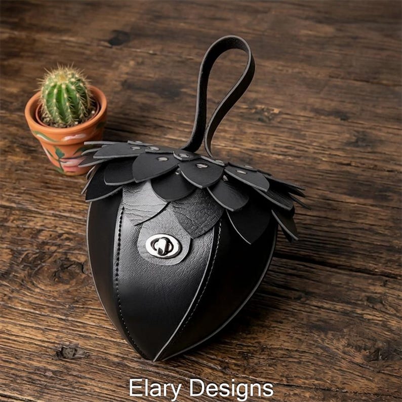 May include: Black leather handbag with a unique acorn design. The bag features a petal-like top, a handle, and a silver clasp. The words "Elary Designs" are printed at the bottom of the image. A small cactus in a terracotta pot is in the background.
