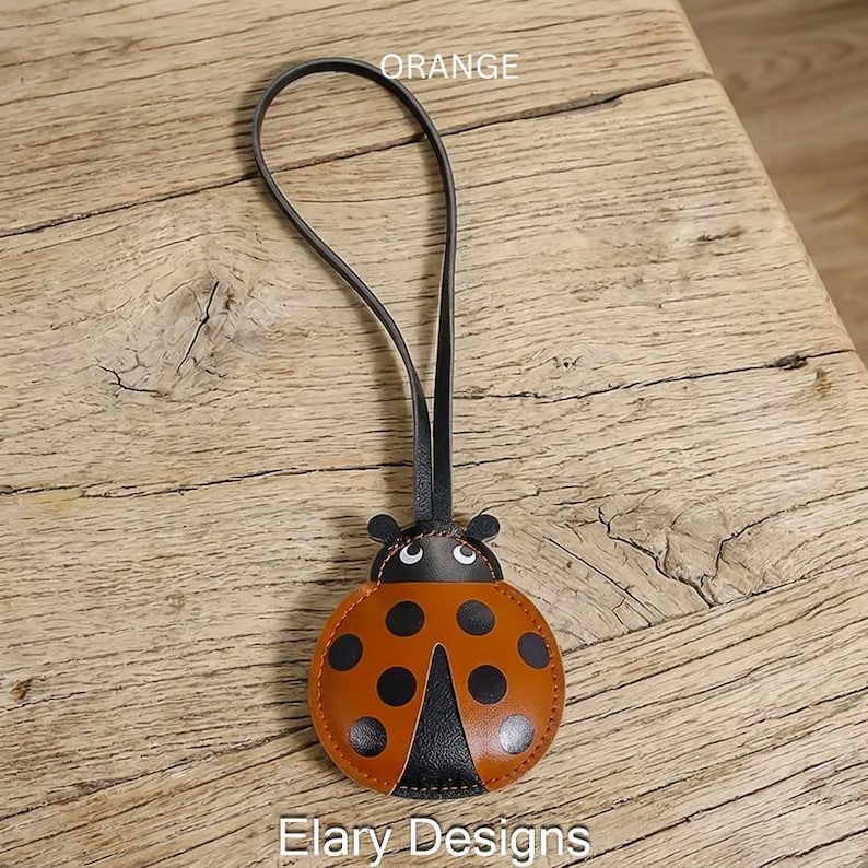 May include: A ladybird-shaped leather charm with a long black strap. The charm features an orange body with black circular spots, a black head section with white eyes, and the text ORANGE and Elary Designs printed on the image.