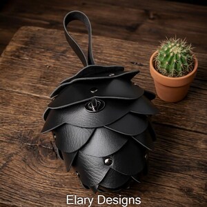May include: Black leather pouch shaped like a pinecone, with a loop handle. The pouch is made of overlapping, petal-like pieces and secured with metal rivets and a clasp. A small cactus in a terracotta pot is in the background. The text "Elary Designs" is at the bottom.