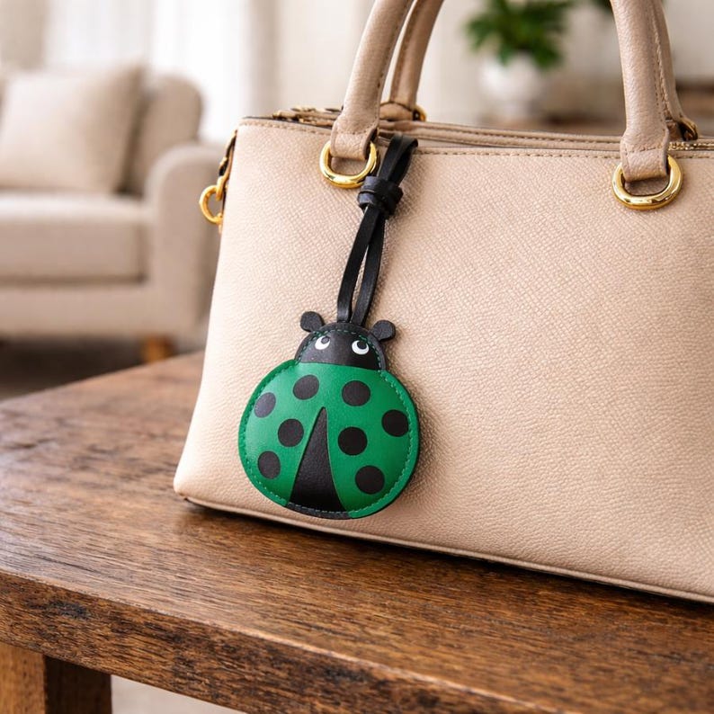 May include: A green and black ladybug-shaped bag charm with black accents hangs from a beige handbag. The ladybug charm has black spots and a black leather strap. The handbag has gold-colored hardware and a top handle.
