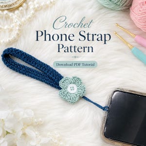 Könnte beinhalten: Eine marineblaue gehäkelte Handyschlaufe mit einer hellgrünen Blume und weißen Knöpfen. Der Riemen ist an einem Telefon befestigt. Das Bild enthält den Text "Crochet Phone Strap Pattern" und "Download PDF Tutorial".