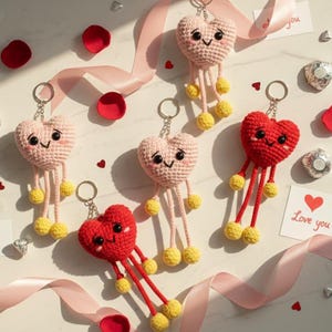 Può includere: Portachiavi a forma di cuore all'uncinetto in rosa e rosso, ciascuno con gambe pendenti e pompon gialli. I portachiavi hanno facce sorridenti e sono disposti con petali di rosa rossi e un nastro. Una carta dice "Love you."