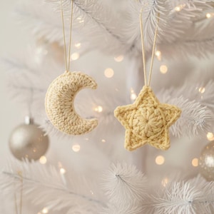 Könnte beinhalten: Zwei handgefertigte Weihnachtsornamente aus Häkelarbeit: ein Halbmond und ein fünfzackiger Stern. Beide Ornamente sind hellbeige und hängen an einem weißen Weihnachtsbaum. Die Ornamente sind an dünnen, beigefarbenen Schnüren aufgehängt.