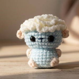 Könnte beinhalten: Eine handgefertigte Häkellammfigur. Das Lamm hat einen hellblauen Körper, weiße, flauschige Wolle und kleine rosa Ohren und Füße. Es hat große schwarze Augen und einen kleinen blau gestickten Mund. Das Lamm sitzt auf einer Holzoberfläche.