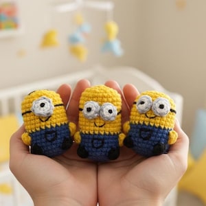 Könnte beinhalten: Drei handgefertigte Minion-Figuren aus Häkelarbeit. Jede Figur ist gelb mit blauen Latzhosen, schwarzen Schuhen und grauen Brillen. Die Minions werden in geformten Händen gehalten, mit einem unscharfen Hintergrund aus einer Krippe und einem Mobile.