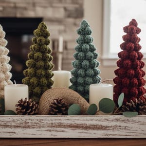 Könnte beinhalten: Eine dekorative Anordnung mit handgefertigten gehäkelten Weihnachtsbäumen in Creme, Grün und Burgunderrot. Die Bäume sind mit weißen Kerzen, Tannenzapfen und Grün in einem rustikalen Holztablett angeordnet.