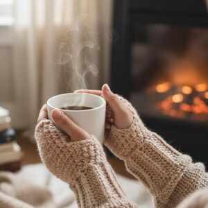 Könnte beinhalten: Eine Person hält eine weiße Keramiktasse mit einem dunklen Getränk, aus dem Dampf aufsteigt. Die Person trägt beige gestrickte Fingerhandschuhe. Im Hintergrund befindet sich ein Kamin mit einem warmen Feuer.