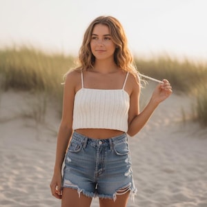 Könnte beinhalten: Ein weißes gehäkeltes Crop-Top mit dünnen Trägern und einem quadratischen Ausschnitt. Das Top wird mit Jeansshorts mit Distressed-Details kombiniert. Das Bild wird im Freien aufgenommen, mit einem Sandstrand und hohem Gras im Hintergrund.