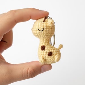 Könnte beinhalten: Ein gehäkeltes Giraffen-Schlüsselanhänger in Hellgelb mit braunen Flecken und einem lächelnden Gesicht. Der Schlüsselanhänger hat einen silbernen Metallring. Die Giraffe ist etwa 7.6 cm groß.