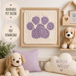 Könnte beinhalten: Ein quadratischer Häkel-Wandbehang mit einem violetten Pfotenabdruck auf cremefarbenem Grund, gerahmt in hellem Holz. Das Bild enthält den Text ADORABLE PET DECOR und PDF DOWNLOAD sowie ein Welpen-Stofftier und ein wolkenförmiges Kissen.
