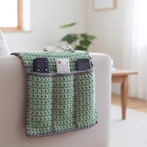 Könnte beinhalten: Ein grüner gehäkelter Sofa-Arm-Organizer mit drei Taschen, grau eingefasst. Der Organizer enthält eine Fernbedienung, eine TV-Fernbedienung und ein Smartphone. Eine Brille liegt oben auf dem Organizer.