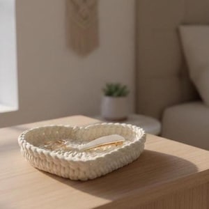 May include: A heart-shaped, off-white woven tray sits on a light wood surface. The tray contains a few gold-colored hair accessories. The scene is set in a bedroom with a window and a bed in the background.