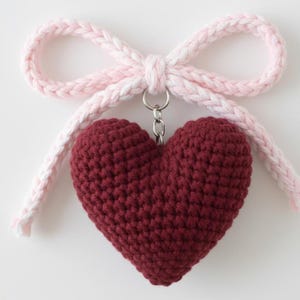 Könnte beinhalten: Ein weinrotes, gehäkeltes herzförmiges Charm mit einer silberfarbenen Kette und einer rosa-weiß geflochtenen Schleife. Das Herz ist einfarbig und hat eine strukturierte Oberfläche. Die Schleife ist oben am Herzen befestigt.