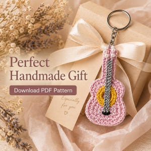 Könnte beinhalten: Ein rosa gehäkelter Gitarren-Schlüsselanhänger mit gelbem Schallloch und silbernen Saiten hängt in der Nähe einer Geschenkbox, die mit einem cremefarbenen Band gebunden ist. Das Bild enthält den Text "Perfect Handmade Gift" und "Download PDF Pattern".