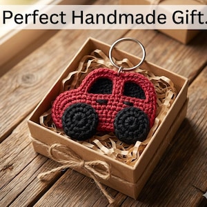 Könnte beinhalten: Ein handgefertigter roter Häkel-Auto-Schlüsselanhänger mit schwarzen Rädern, präsentiert in einer kleinen Geschenkbox mit Papierfüllung. Die Box ist mit Bindfaden gebunden. Der Text "Perfect Handmade Gift" steht oben.