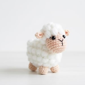 May include: A crocheted sheep toy with a fluffy white body, a light pink face, ears, and feet, and black eyes. The toy is standing on a white surface.