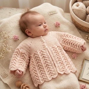 Könnte beinhalten: Ein Baby in einem hellrosa gehäkelten Cardigan mit einem dekorativen Blättermuster und einem gewellten Saum. Das Baby liegt auf einer cremefarbenen Decke, die mit rosa Rosen verziert ist. Ein Korb mit Garn und ein gerahmtes Bild befinden sich im Hintergrund.