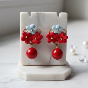 Könnte beinhalten: Handgefertigte Ohrringe mit roten und hellblauen floralen Designs. Jeder Ohrring hat eine Ansammlung roter Blumen, hellblauer Blumen und eine hängende rote Kugel. Die Ohrringe werden auf einem weißen Marmorständer präsentiert.