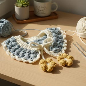 Può includere: Un colletto all'uncinetto fatto a mano in una delicata combinazione di colori blu e crema, con un design a punto puff testurizzato. Il colletto è adornato da un fiocco color crema e due piccoli fiori all'uncinetto gialli. Sono visibili anche filati e uncinetti.