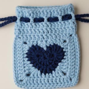 May include: A light blue crochet drawstring bag with a dark blue heart design in the center. The bag has a square shape with a textured pattern and dark blue drawstring cords.