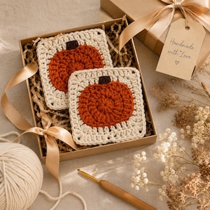 Könnte beinhalten: Zwei quadratische Häkeluntersetzer mit einem zentralen orangefarbenen Kürbismotiv und einem dunkelbraunen Stiel auf cremefarbenem Hintergrund. Die Untersetzer befinden sich in einer Geschenkbox aus Karton mit beigem Band und einem Etikett.