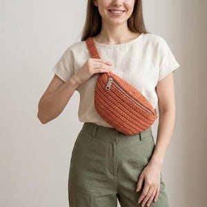Könnte beinhalten: Eine orangefarbene Häkeltasche mit Reißverschluss. Die Tasche wird quer über dem Körper getragen. Die Tasche besteht aus einem strukturierten, gewebten Material. Die Tasche hat eine gebrannte orange Farbe.