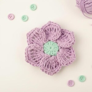 Könnte beinhalten: Eine handgefertigte, lavendelfarbene Häkelblume mit einem mintgrünen Zentrum. Kleine, pastellfarbene Knöpfe in Lavendel und Mintgrün umgeben die Blume. Ein Knäuel Lavendelgarn deutet auf Bastelarbeiten hin.
