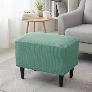 May include: A rectangular ottoman with a teal, textured fabric cover and four black legs. The ottoman is in a living room setting, with a small side table, a gray armchair, and a white bookshelf in the background.