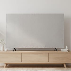 Puede incluir: Una funda de TV gris claro cubre un televisor de pantalla plana. El televisor está sobre un mueble de madera clara con tres cajones. Una planta en maceta, una lámpara pequeña y objetos decorativos están sobre el mueble. La habitación tiene una paleta de colores neutros.