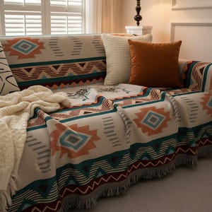 Könnte beinhalten: Eine cremefarbene Sofadecke mit einem geometrischen Muster in Türkis, Orange und Braun. Die Decke hat einen Fransenrand und ist über einem Sofa mit passenden Wurfkissen drapiert. Eine cremefarbene Strickdecke ist ebenfalls vorhanden.