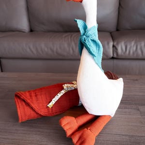 May include: A plush goose toy with a white body, orange beak and feet, and a teal scarf. A folded red blanket with a floral ribbon is next to the goose. The toy is on a wooden surface.