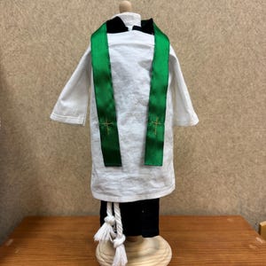 Puede incluir: Una camisa de clérigo blanca en miniatura con una estola verde con cruces doradas. El atuendo incluye pantalones negros y una cuerda blanca con borlas. El conjunto se exhibe sobre un soporte de madera.