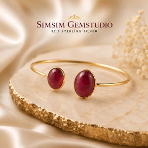 May include: A gold-toned open cuff bracelet featuring two oval-shaped deep red gemstones at each end. The text SIMSIM GEMSTUDIO 92.5 STERLING SILVER is visible above the jewellery. The bracelet has a smooth, polished metal band and setting.