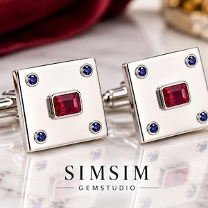 Puede incluir: Un par de gemelos cuadrados de tono plateado con una piedra preciosa roja rectangular central rodeada por cuatro pequeñas piedras azules circulares en las esquinas. El texto SIMSIM GEMSTUDIO está impreso en negro debajo de los gemelos.