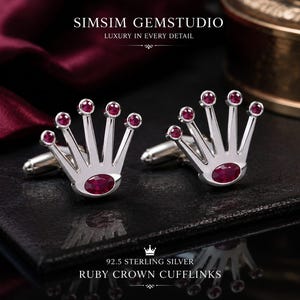 May include: A pair of silver-toned cufflinks shaped like a crown, each featuring five small round red rubies at the tips and one larger oval red ruby at the base. The text SIMSIM GEMSTUDIO, 92.5 STERLING SILVER, and RUBY CROWN CUFFLINKS is visible.