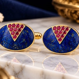 May include: A pair of oval-shaped gold cufflinks featuring a deep blue lapis lazuli inlay. Each cufflink is accented with a triangular cluster of ten small, round, vibrant red rubies set in gold, positioned at the top center of the blue stone.