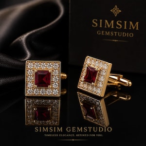 May include: A pair of gold-toned square cufflinks featuring a central princess-cut deep red gemstone surrounded by a border of small clear round stones. The cufflinks are displayed near a black box with the text SIMSIM GEMSTUDIO printed on it.
