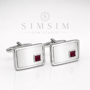 Puede incluir: Un par de gemelos plateados con diseño rectangular. Cada gemelo presenta una superficie plateada pulida y una gema cuadrada de color rojo intenso en el centro. Los gemelos se muestran sobre un fondo blanco. El texto "SIMSIM GEM STUDIO" está en la parte superior.